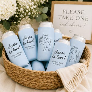 K&ouml;nnte beinhalten: Eine Sammlung hellblauer Dosenk&uuml;hler aus Schaumstoff in einem geflochtenen Korb. Einige K&uuml;hler tragen den schwarzen Text "cheers to love!" mit "MIRANDA & CHASE 06.20.2026" darunter, andere zeigen eine schwarze Strichzeichnung einer Katze mit Flasche.