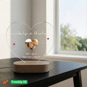 Puede incluir: Una placa de acrílico transparente en forma de corazón con los nombres Emily y Charlie, una foto central en forma de corazón de una pareja, dos contornos de mapas con corazones rojos y el texto Miles apart, but close at heart, sobre una base de madera.