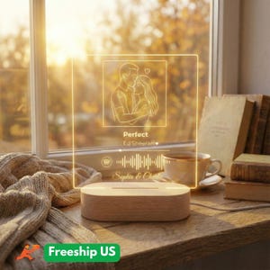 Può includere: Lampada a LED con base in legno e pannello in acrilico trasparente con un disegno a tratto di una coppia che si abbraccia, il titolo della canzone "Perfect" di Ed Sheeran e i nomi "Sophia & Charlie". La lampada è su una superficie di legno.