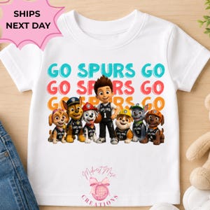 May include: A white short-sleeve t-shirt featuring a graphic of six Paw Patrol characters wearing San Antonio Spurs basketball jerseys. Above the characters, the text GO SPURS GO is printed in three rows of teal, red, and orange colors.