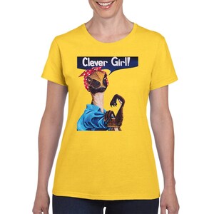 以下が含まれることがあります： 「Clever Girl!」というテキストと、赤と白のドット柄のバンドゥと青いシャツを着た恐竜のグラフィックが描かれた黄色のTシャツ