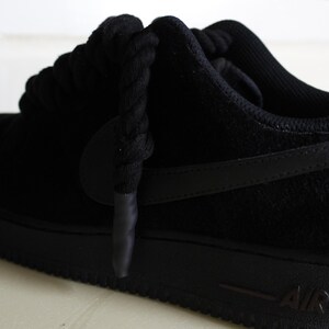 May include: Black Air Force 1 sneakers. The shoe features a black suede upper, a black sole, and black laces. The iconic Nike swoosh is visible on the side. The word "AIR" is printed on the side of the sole.