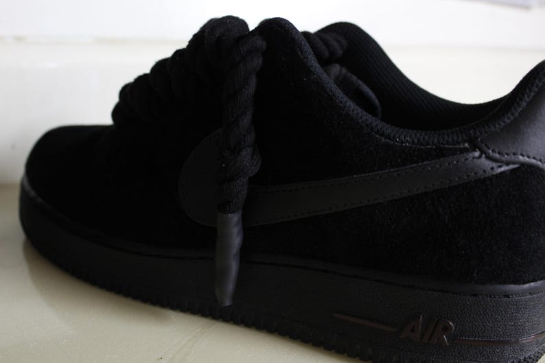 May include: Black Air Force 1 sneakers. The shoe features a black suede upper, a black sole, and black laces. The iconic Nike swoosh is visible on the side. The word "AIR" is printed on the side of the sole.