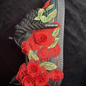 May include: Black sneaker with intricate red rose embroidery and green leaves. The floral design adorns the side and toe of the shoe, creating a striking contrast against the black background. The laces are black.