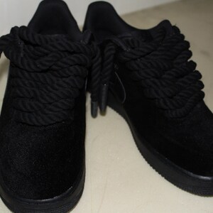May include: Pair of black sneakers with thick, rope-like laces. The shoes have a low profile and a solid black sole. The laces are wrapped around the top of the shoe, creating a unique design. The material appears to be a soft, suede-like fabric.