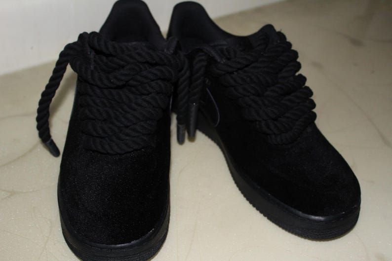 May include: Pair of black sneakers with thick, rope-like laces. The shoes have a low profile and a solid black sole. The laces are wrapped around the top of the shoe, creating a unique design. The material appears to be a soft, suede-like fabric.