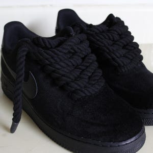 May include: A pair of black sneakers with thick, rope-like laces. The shoes have a suede-like texture and a black sole. The Nike swoosh logo is visible on the side. The overall style is modern and urban.