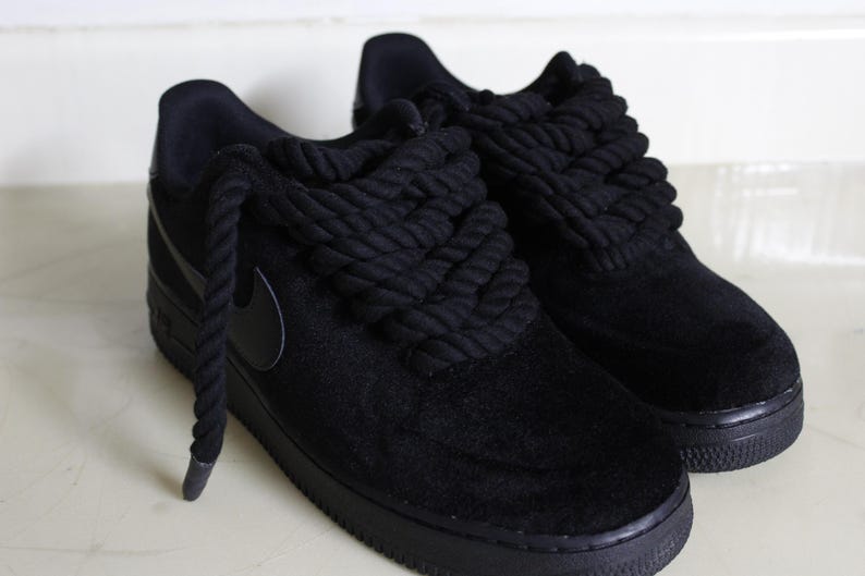 May include: A pair of black sneakers with thick, rope-like laces. The shoes have a suede-like texture and a black sole. The Nike swoosh logo is visible on the side. The overall style is modern and urban.