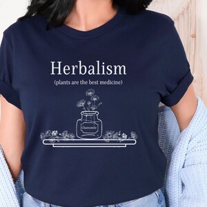 May include: Navy blue t-shirt with white text that reads "Herbalism (plants are the best medicine)". A white line drawing of a jar with flowers and the word "Chamomile" is on the shirt.
