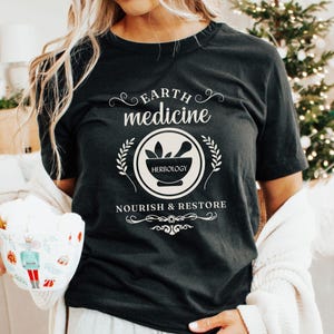 May include: Black t-shirt with the text "Earth Medicine" and a circular design featuring a mortar and pestle, with the word "Herbology" inside. The shirt also has the words "Nourish & Restore". A festive mug with a red and white striped straw is held in the image.