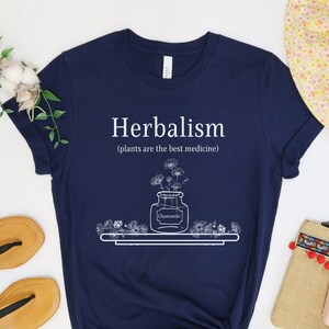 May include: Navy blue t-shirt with white text that reads "Herbalism (plants are the best medicine)". The design features a jar of chamomile flowers on a shelf.