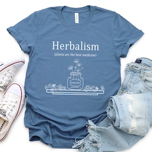 May include: A blue t-shirt with white text that reads "Herbalism (plants are the best medicine)". The shirt features a white illustration of a jar of chamomile flowers.