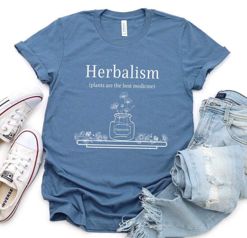 May include: A blue t-shirt with white text that reads "Herbalism (plants are the best medicine)". The shirt features a white illustration of a jar of chamomile flowers.