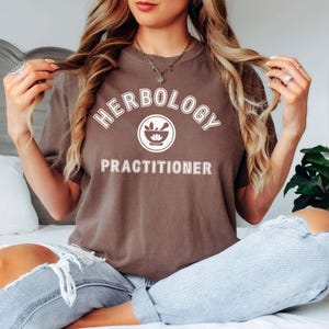 May include: A brown t-shirt with the words "HERBOLOGY PRACTITIONER" in white, arched above a circular graphic of a mortar and pestle. The shirt is a relaxed fit, and the model is wearing light-wash denim jeans.