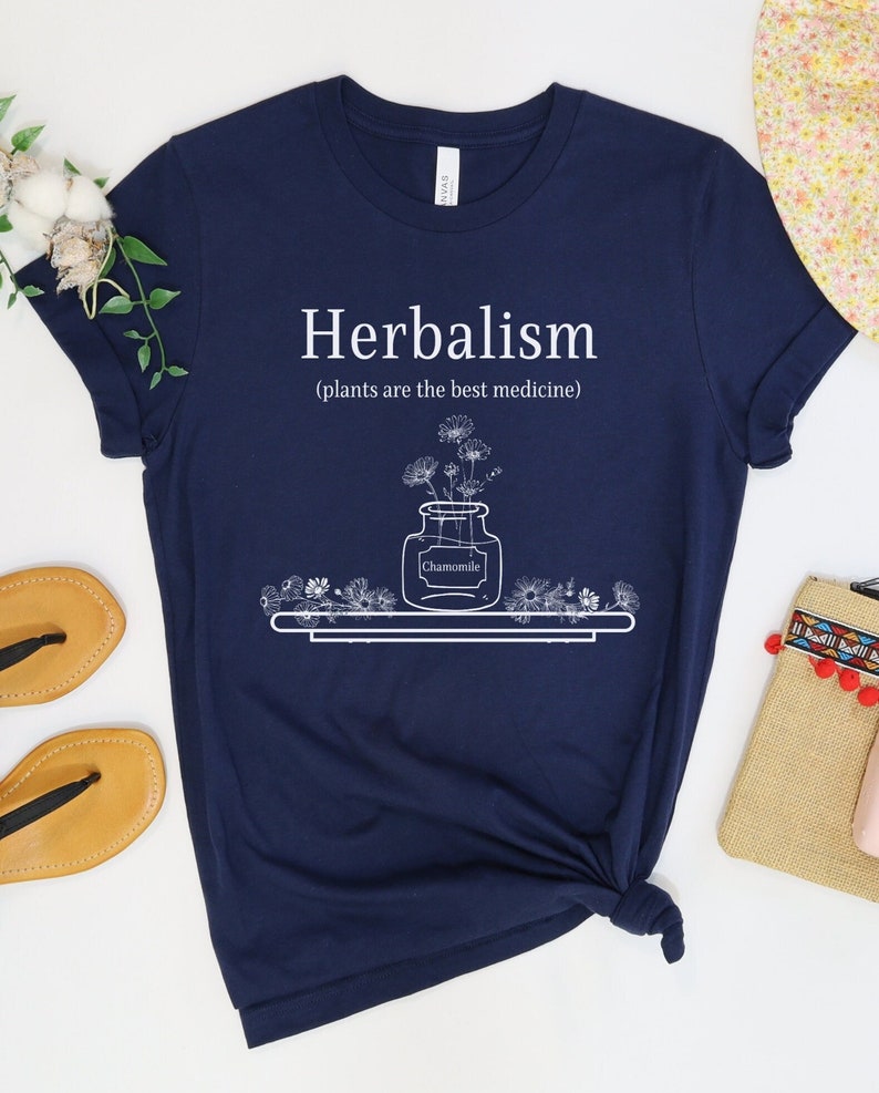 May include: Navy blue t-shirt with white text that reads "Herbalism (plants are the best medicine)". The design features a jar of chamomile flowers on a shelf.