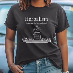 May include: A black t-shirt with white text that reads "Herbalism (plants are the best medicine)". A drawing of a jar with flowers and the word "Chamomile" is on the shirt.