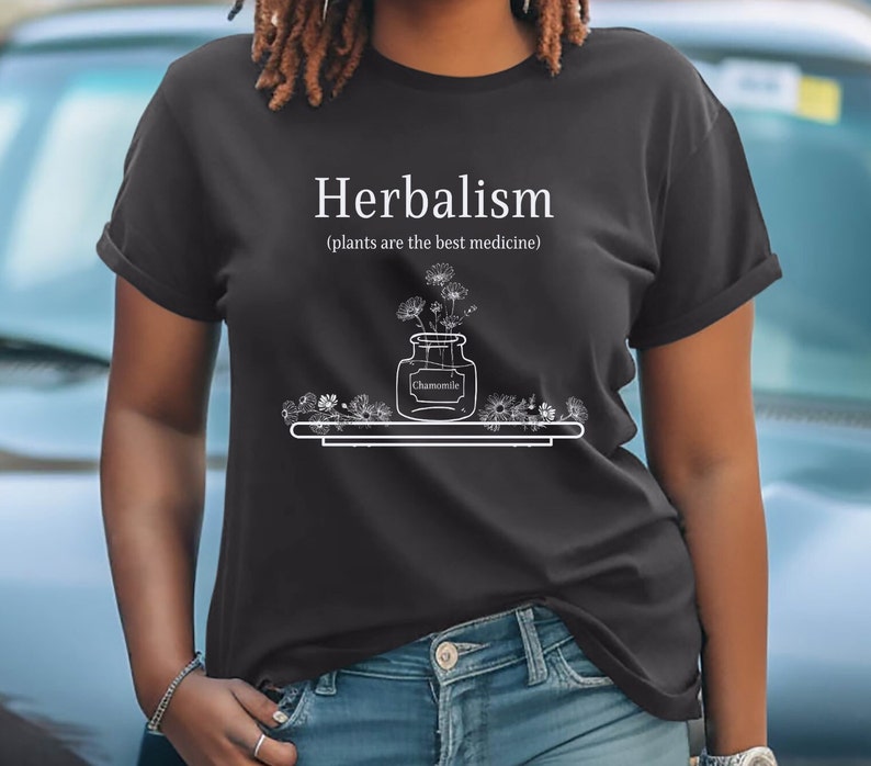 May include: A black t-shirt with white text that reads "Herbalism (plants are the best medicine)". A drawing of a jar with flowers and the word "Chamomile" is on the shirt.
