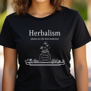 May include: Black t-shirt with white text that reads "Herbalism (plants are the best medicine)". A drawing of a jar with flowers and the word "Chamomile" is below the text.