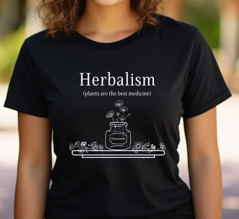 May include: Black t-shirt with white text that reads "Herbalism (plants are the best medicine)". A drawing of a jar with flowers and the word "Chamomile" is below the text.