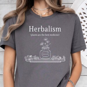 May include: A gray t-shirt with the text "Herbalism (plants are the best medicine)" and a drawing of a jar with flowers and the word "Chamomile" on a label.