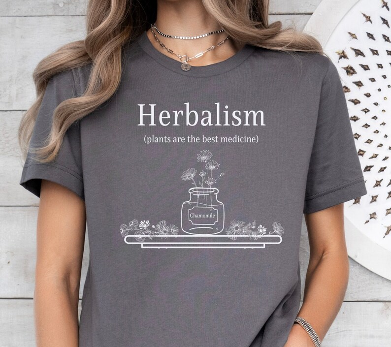 May include: A gray t-shirt with the text "Herbalism (plants are the best medicine)" and a drawing of a jar with flowers and the word "Chamomile" on a label.