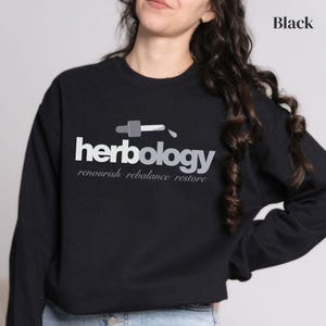 Könnte beinhalten: Schwarzes Sweatshirt mit dem Wort "herbology" in Weiß und Grau sowie einer Tropfer-Grafik. Darunter stehen die Worte "renourish rebalance restore" in Grau. Das Wort "Black" befindet sich in der oberen rechten Ecke.