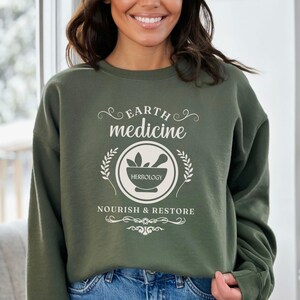 May include: Olive green sweatshirt with the text "Earth Medicine" and "Herbology" in white. The design includes a mortar and pestle inside a circle, surrounded by laurel branches, and the words "Nourish & Restore".