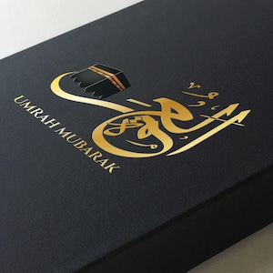 May include: A black box with gold lettering that says "Umrah Mubarak" and a gold illustration of the Kaaba.