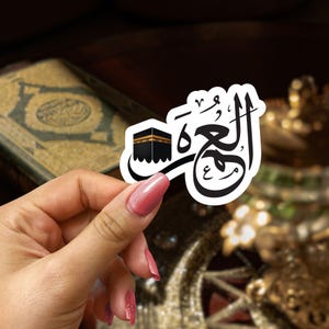 May include: A white sticker with black Arabic calligraphy that says "Hajj" and a black silhouette of the Kaaba.