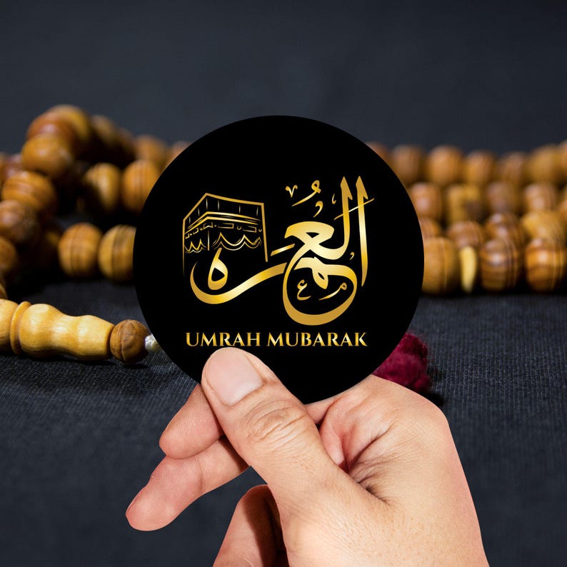 May include: A black circle with gold calligraphy and a small image of the Kaaba. The text reads "Umrah Mubarak".