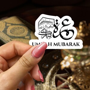 May include: White sticker with black calligraphy and a drawing of the Kaaba. The text reads "Umrah Mubarak".