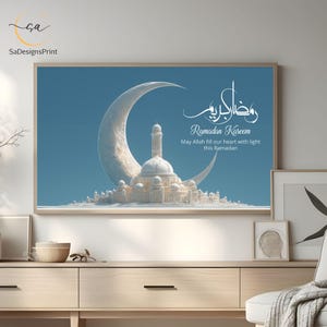 Pu&ograve; includere: Una stampa incorniciata raffigurante una moschea bianca e una luna crescente su uno sfondo blu. La stampa include il testo "Ramadan Kareem" e un messaggio. L'opera d'arte &egrave; esposta su un mobile in legno chiaro.