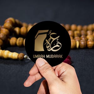 May include: A black round sticker with gold Arabic calligraphy and the text "Umrah Mubarak". The sticker is being held by a hand against a background of brown wooden beads.