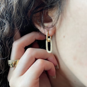 Peut inclure: Boucles d'oreilles dorées en forme de chaîne avec un pendentif rectangulaire orné de cristaux. Les boucles d'oreilles sont portées à l'oreille, et une bague dorée avec des cristaux est au doigt. Les bijoux sont présentés sur un fond flou.