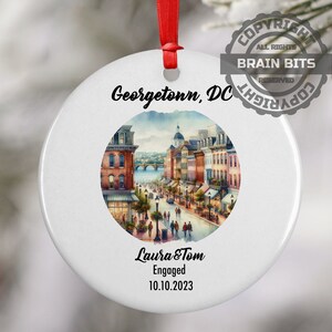 Peut inclure: Ornement en céramique blanche avec une illustration aquarelle de Georgetown, DC. Le texte "Georgetown, DC" est imprimé au-dessus de l'illustration. Sous l'illustration, le texte "Laura & Tom Engagés 10.10.2023" est imprimé.