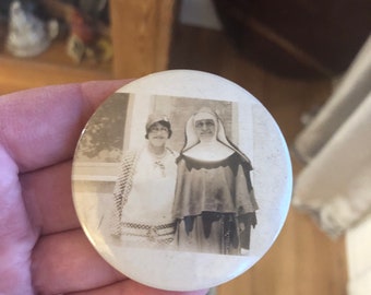 May include: A vintage black and white photo of two women, one wearing a hat and the other wearing a nun's habit, is displayed on a round button.