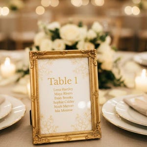 Puede incluir: Una tarjeta dorada con el número de mesa "Mesa 1" y los nombres de los invitados. La tarjeta tiene un diseño floral decorativo y está colocada sobre una mesa con platos blancos y velas, lo que sugiere una boda o evento formal.