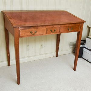 May include: A wooden writing desk with a brown leather top and three drawers. The desk has a simple design with straight legs and a rectangular top.
