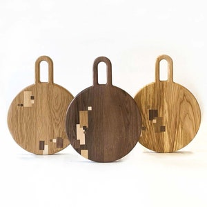 Puede incluir: Tres tablas de cortar redondas de madera con asas. Las tablas están hechas de diferentes tipos de madera, incluyendo nogal y roble. Las tablas tienen un patrón geométrico de cuadrados incrustados en la superficie.