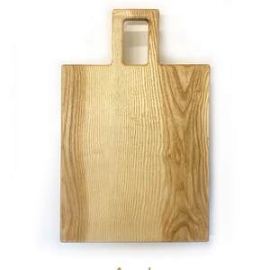 May include: A rectangular wooden chopping board with a handle. The board is made of ash wood and has a natural finish. The handle is a square cutout with a smaller square cutout in the centre. The board is labelled "Ash".