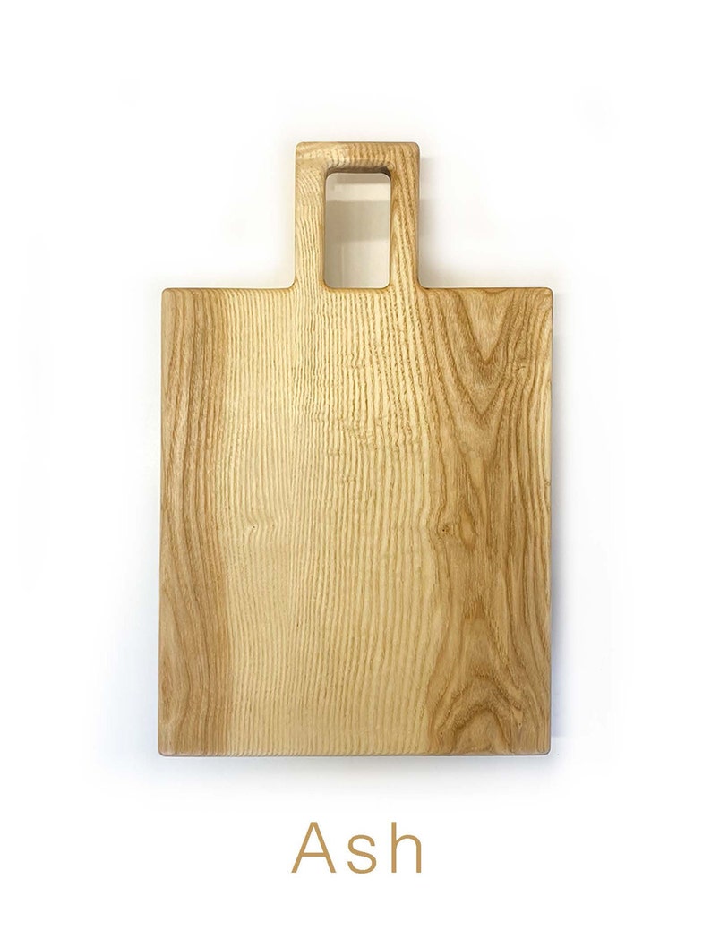 May include: A rectangular wooden chopping board with a handle. The board is made of ash wood and has a natural finish. The handle is a square cutout with a smaller square cutout in the centre. The board is labelled "Ash".