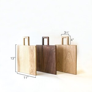 May include: Three wooden chopping boards with handles. The boards are 13 inches tall, 11 inches wide, and 4 inches deep. The boards are made of different types of wood, including light wood and dark wood.