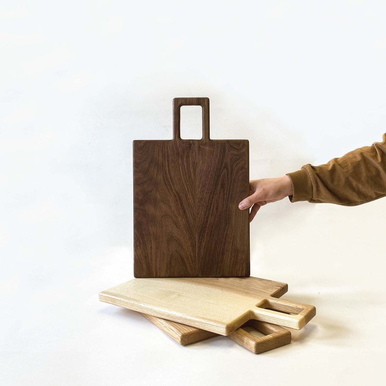 May include: Three wooden chopping boards, one dark brown with a handle and two lighter wood boards with handles. The boards are stacked on top of each other.