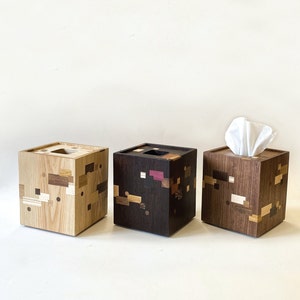 Può includere: Tre porta fazzoletti in legno con diversi motivi in legno. La scatola a sinistra è in legno chiaro con accenti marroni e bianchi. La scatola al centro è in legno scuro con accenti marroni e bianchi. La scatola a destra è in legno scuro con accenti marroni e bianchi e contiene un fazzoletto bianco.