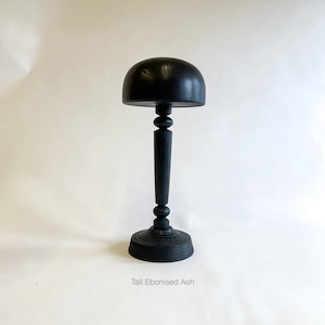 Op de afbeelding: Een hoge zwarte houten hoedenstandaard met een ronde bovenkant en een voet. De standaard is gemaakt van ge&euml;boniseerde essenhout. "Tall Ebonised Ash" staat onder de standaard.