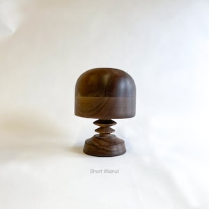 Op de afbeelding: Een korte pruikenstandaard van walnoot hout met een afgeronde bovenkant en een getrapte basis. "Short Walnut" staat onder de standaard.