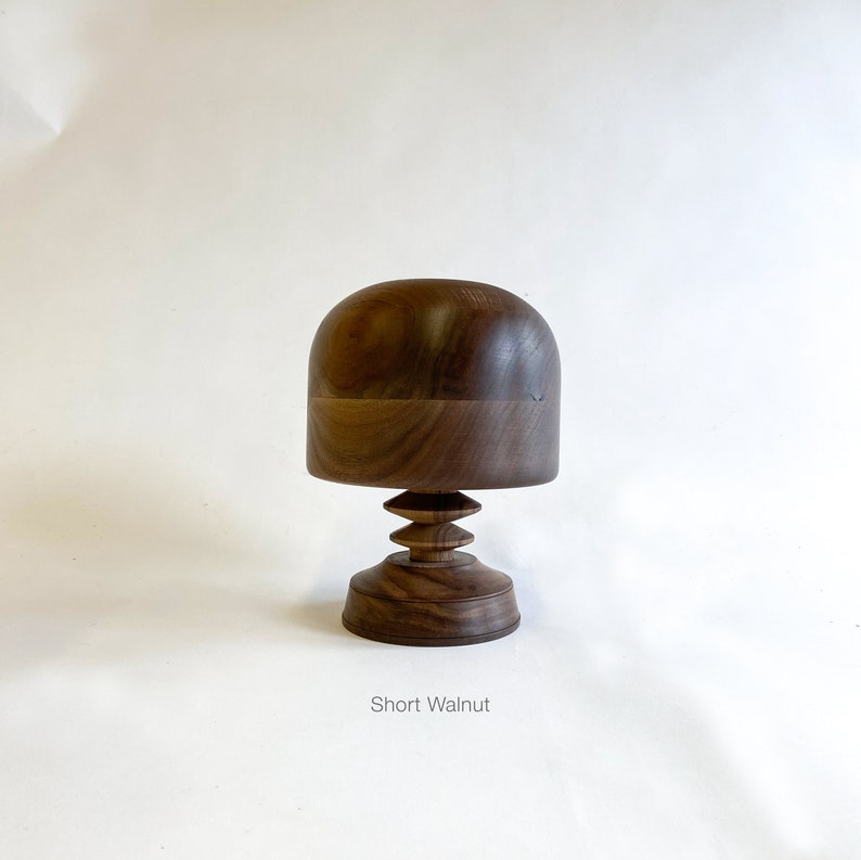 Op de afbeelding: Een korte pruikenstandaard van walnoot hout met een afgeronde bovenkant en een getrapte basis. "Short Walnut" staat onder de standaard.