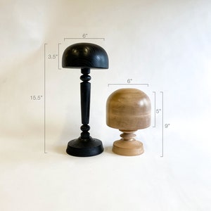 Op de afbeelding: Twee houten hoedvormen, een zwarte en een van naturel hout. De zwarte hoedvorm is 15,2 cm hoog en 8,9 cm breed. De hoedvorm van naturel hout is 15,2 cm hoog en 12,7 cm breed. Beide hoedvormen staan op een witte achtergrond.