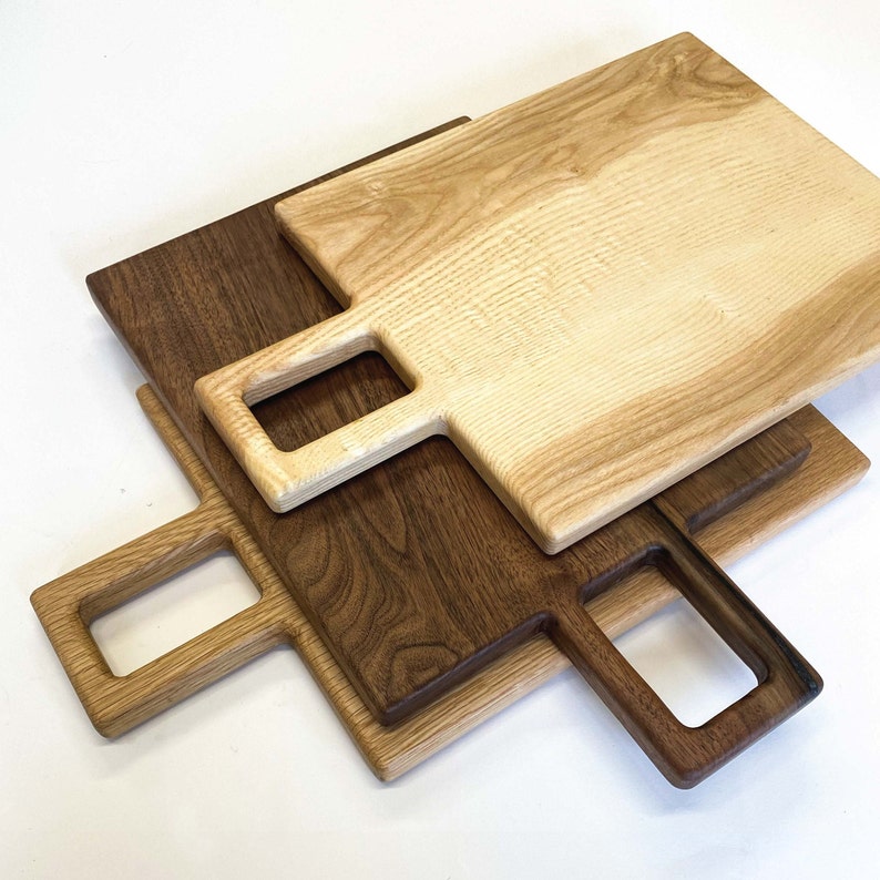 May include: Three wooden chopping boards with handles. The top board is light wood, the middle board is dark wood, and the bottom board is light wood. The boards are stacked on top of each other.