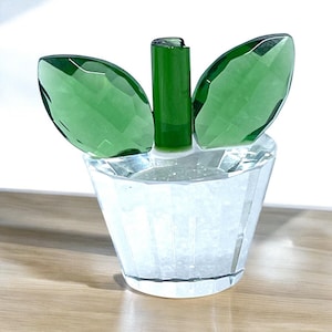 May include: A clear crystal flower pot with green leaves and stem. The leaves are faceted and angled, and the pot has a geometric design. The flower pot is sitting on a wooden surface.